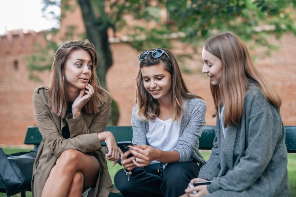 Tre giovani donne sono sedute sulla panchina e utilizzano il telefono cellulare. P — Foto Stock