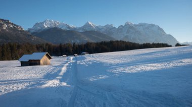Karwendel dağları manzaralı ahşap kulübelerle kaplı karla kaplı Buckelwiesen çayırı. Boşluğu kopyalayan mavi gökyüzü