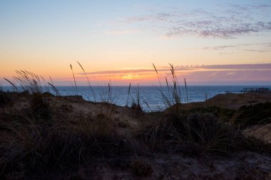 Deniz kenarında gün batımı, batan güneş, renkli gökyüzü ve kumullardaki deniz otları, Portekiz kıyıları.