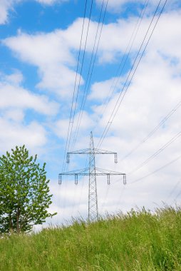 one power pole with high-voltage wires on grassy hill, tree beside. blue sky with clouds. vertical format.