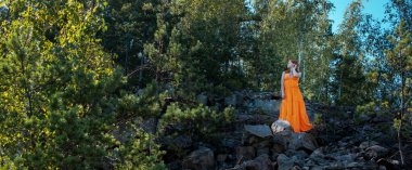 Parlak turuncu elbiseli bir kadın yaprakların arasında kayalık bir yamaçta duruyor ve vahşi doğada güneşli bir yaz gününün tadını çıkarıyor. Panoramik fotoğraf.