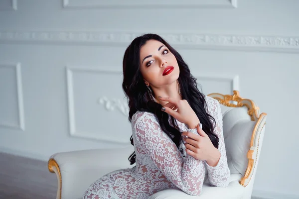 Portrait of perfect, sexy young woman with long black hair and red lips wearing seductive white dress sitting on luxury vintage chair. — Stock fotografie