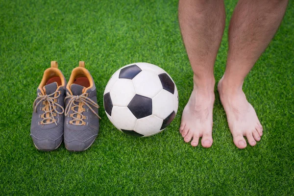 Pernas de jogador de futebol descalço irreconhecível, bola de futebol e sapatos esportivos contra grama artificial. — Fotografia de Stock