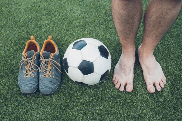 Pernas de jogador de futebol descalço irreconhecível, bola de futebol e sapatos esportivos contra grama artificial. — Fotografia de Stock