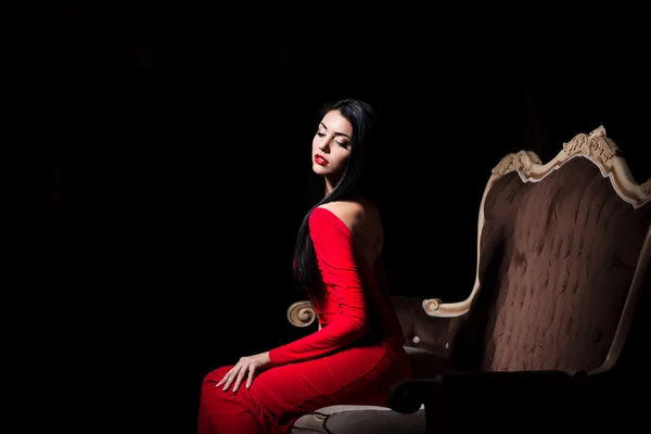 Sensual female in red dress posing in a sexy way on luxury sofa in dramatic light — Stock fotografie