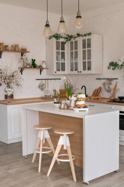 Paskalya pastaları, çiçekler, yumurtalar ve masanın üstünde şenlikli bir dekorla aydınlık bir bahar mutfağı sahnesi. Doğal ışık ve yumuşak renkler sıcak mevsimlik tatil atmosferi yaratır..