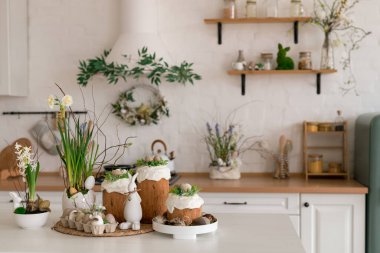 Paskalya pastaları, çiçekler, yumurtalar ve masanın üstünde şenlikli bir dekorla aydınlık bir bahar mutfağı sahnesi. Doğal ışık ve yumuşak renkler sıcak mevsimlik tatil atmosferi yaratır..