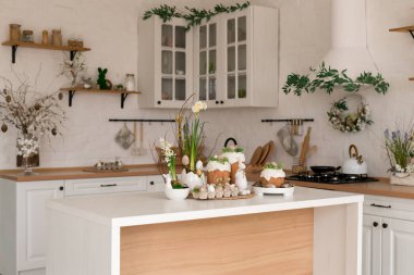 Paskalya pastaları, çiçekler, yumurtalar ve masanın üstünde şenlikli bir dekorla aydınlık bir bahar mutfağı sahnesi. Doğal ışık ve yumuşak renkler sıcak mevsimlik tatil atmosferi yaratır..