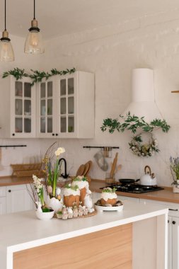 Paskalya pastaları, çiçekler, yumurtalar ve masanın üstünde şenlikli bir dekorla aydınlık bir bahar mutfağı sahnesi. Doğal ışık ve yumuşak renkler sıcak mevsimlik tatil atmosferi yaratır..