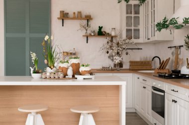 Paskalya pastaları, çiçekler, yumurtalar ve masanın üstünde şenlikli bir dekorla aydınlık bir bahar mutfağı sahnesi. Doğal ışık ve yumuşak renkler sıcak mevsimlik tatil atmosferi yaratır..