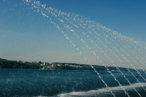 Jets of water from the fountain overflow in the sun and fall into the lake. In the background of the photo you can see the lake and houses.