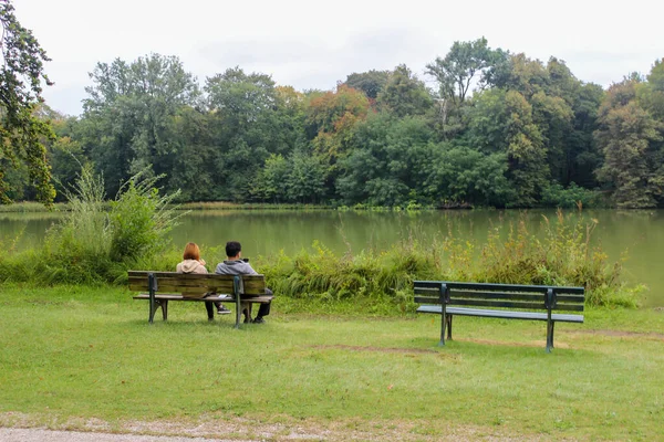 城市公园池塘岸边的两张长椅的壮丽景色 在一张长椅上坐着两个年轻人 — 图库照片