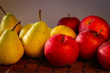 Olgun sarı armutlar ve kırmızı elmalar güneşin ışınlarıyla aydınlatılmış hasır bir sepetin üzerinde duruyor.