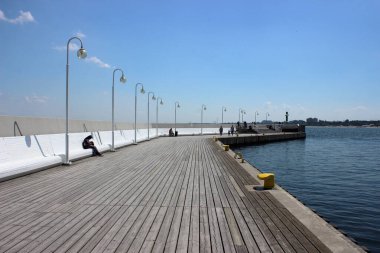 Açık güneşli bir günde, deniz kenarında beyaz bankları ve sokak lambaları olan geniş ahşap gezinti güvertesi. Deniz manzarası seyreden ve eğlenen insanlar.