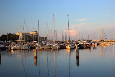 Polonya 'nın Gdynia kentindeki marina manzarası, 5 Haziran 2019, gün batımında. Demirlemiş yelkenli yatlar, açık mavi ve turuncu bir akşam gökyüzünün altındaki limanın sakin sularına yansıyor. Baltık Denizi kıyısında huzurlu bir atmosfer.