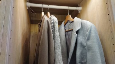 İçi nötr paltolarla dolu minimalist bir gardırop ve modern bir dolabın içindeki yumuşak, sıcak ışıklandırmalı ahşap askılarda asılı örgü giysiler.