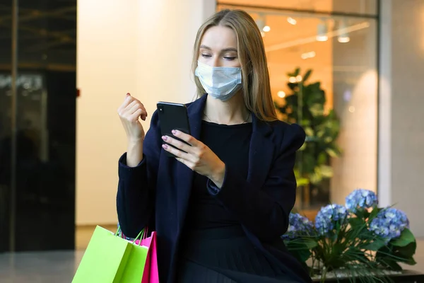 A young woman in a protective mask types on her phone after shopping