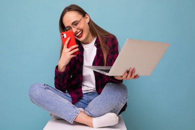 Mavi arka planda dizüstü bilgisayarı ve telefonu olan genç bir kadın oturuyor.