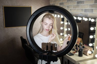 Genç bir kadın yüzük lambasının önünde duruyor ve internetten makyaj kursları veriyor. 