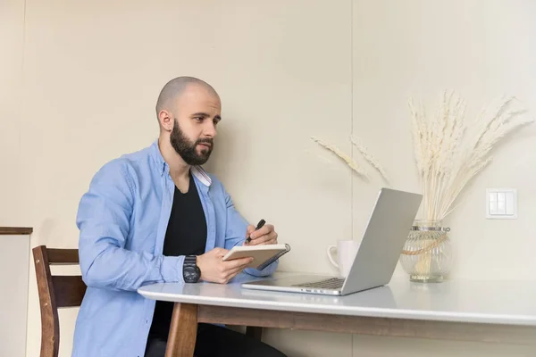 Mavi gömlekli ve siyah kotlu bir adam evde oturmuş bilgisayar ve günlük kullanarak internetten ders çalışıyor.