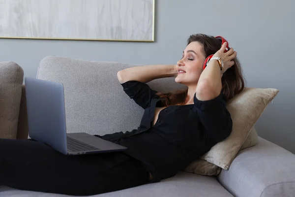 A young woman is lying on the couch listening to music and working on a laptop at home