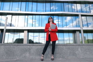 Ceketli bir iş kadını, bir ofis binasının arka planında dizüstü bilgisayarla poz veriyor.