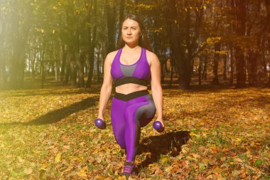 Parlak eşofmanlı bir kız parkta dambıllarla egzersiz yapıyor. Ayarlanmış