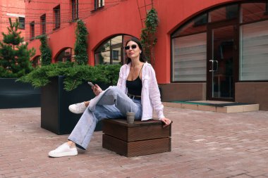 Genç bir kadın elinde bir fincan kahve ve bir cep telefonuyla bankta oturuyor.