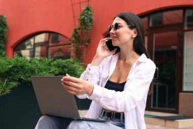 Genç bir kadın bankta oturmuş dizüstü bilgisayarla çalışıyor ve sokakta telefonla konuşuyor.