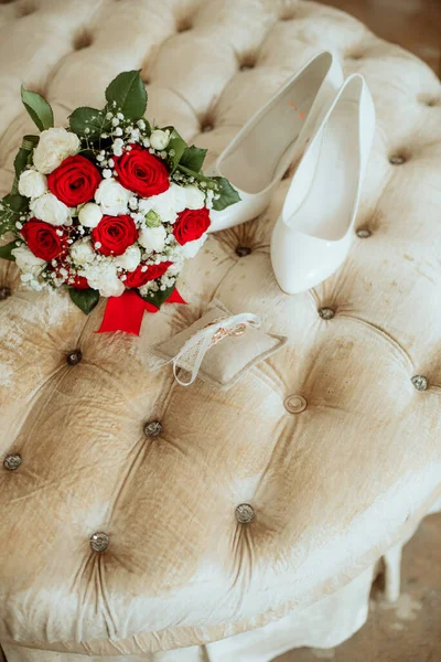 Wedding bouquet of white and red roses, white high heels and gold wedding rings on a boudoir ottoman. Wedding details