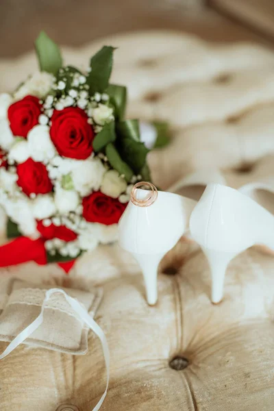 Wedding bouquet of white and red roses, white high heels and gold wedding rings on a boudoir ottoman. Wedding details