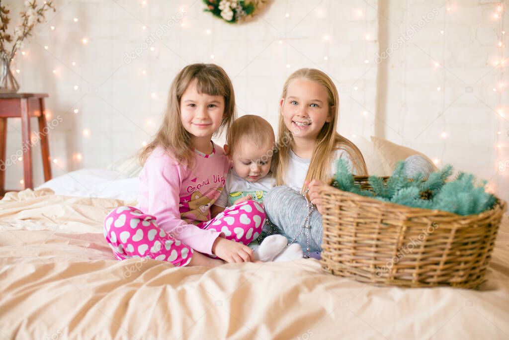 Cute sisters and little brother  in pajamas have fun in in a Scandinavian style bedroom decorated with Christmas garlands and needles on a large bright bed. Christmas mood. Christmas mood. Family time