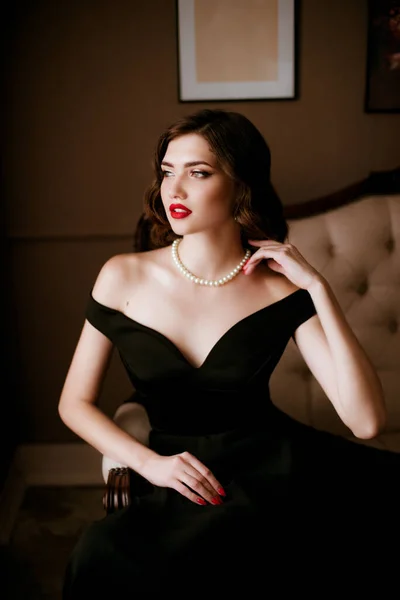 漂亮的年轻女子穿着黑色晚礼服和珍珠项链 穿着可可香奈儿风格的经典化妆品 躺在一个经典的内饰沙发上 美与时尚 — 图库照片