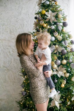 Sarışın, sevimli küçük çocuk, Noel ağacının yanında süslenmiş parlak bir odada altın sarısı parlak elbiseli annesiyle oynuyor. Mutlu bir çocukluk. Noel havası