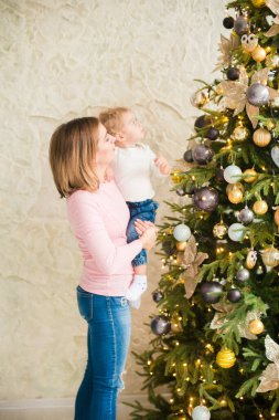 Sarışın, sevimli küçük çocuk, Noel ağacının yanında süslenmiş parlak bir odada annesiyle oynuyor. Mutlu bir çocukluk. Noel havası
