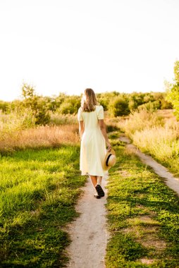 Sarışın, kıvırcık saçlı, açık sarı elbiseli ve elinde hasır bir şapkayla gün batımında kırsaldaki bir tarlada genç bir kız. İnsanlar ve doğa 