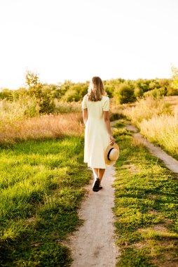 Sarışın, kıvırcık saçlı, açık sarı elbiseli ve elinde hasır bir şapkayla gün batımında kırsaldaki bir tarlada genç bir kız. İnsanlar ve doğa 
