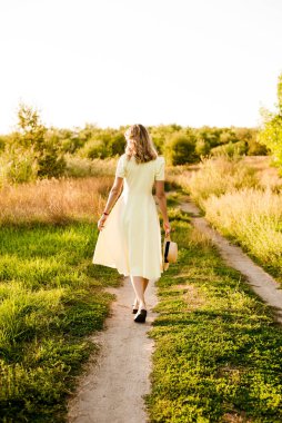 Sarışın, kıvırcık saçlı, açık sarı elbiseli ve elinde hasır bir şapkayla gün batımında kırsaldaki bir tarlada genç bir kız. İnsanlar ve doğa 