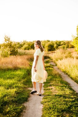 Sarışın, kıvırcık saçlı, açık sarı elbiseli ve elinde hasır bir şapkayla gün batımında kırsaldaki bir tarlada genç bir kız. İnsanlar ve doğa 