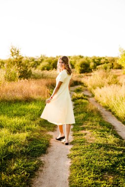Sarışın, kıvırcık saçlı, açık sarı elbiseli ve elinde hasır bir şapkayla gün batımında kırsaldaki bir tarlada genç bir kız. İnsanlar ve doğa 
