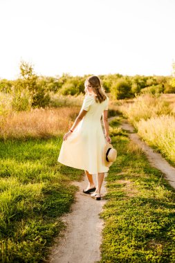 Sarışın, kıvırcık saçlı, açık sarı elbiseli ve elinde hasır bir şapkayla gün batımında kırsaldaki bir tarlada genç bir kız. İnsanlar ve doğa 