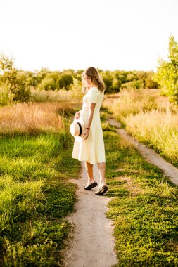 Sarışın, kıvırcık saçlı, açık sarı elbiseli ve elinde hasır bir şapkayla gün batımında kırsaldaki bir tarlada genç bir kız. İnsanlar ve doğa 