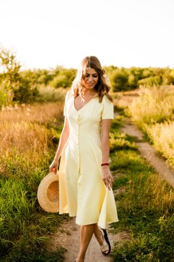 Sarışın, kıvırcık saçlı, açık sarı elbiseli ve elinde hasır bir şapkayla gün batımında kırsaldaki bir tarlada genç bir kız. İnsanlar ve doğa 