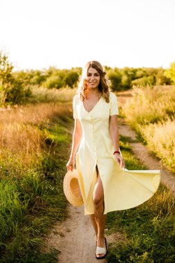 Sarışın, kıvırcık saçlı, açık sarı elbiseli ve elinde hasır bir şapkayla gün batımında kırsaldaki bir tarlada genç bir kız. İnsanlar ve doğa 