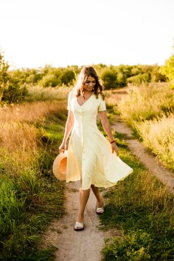 Sarışın, kıvırcık saçlı, açık sarı elbiseli ve elinde hasır bir şapkayla gün batımında kırsaldaki bir tarlada genç bir kız. İnsanlar ve doğa 