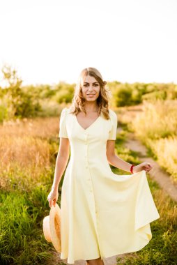 Sarışın, kıvırcık saçlı, açık sarı elbiseli ve elinde hasır bir şapkayla gün batımında kırsaldaki bir tarlada genç bir kız. İnsanlar ve doğa 