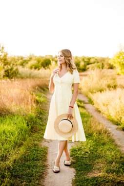 Sarışın, kıvırcık saçlı, açık sarı elbiseli ve elinde hasır bir şapkayla gün batımında kırsaldaki bir tarlada genç bir kız. İnsanlar ve doğa 