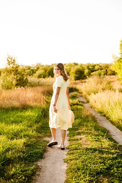 Sarışın, kıvırcık saçlı, açık sarı elbiseli ve elinde hasır bir şapkayla gün batımında kırsaldaki bir tarlada genç bir kız. İnsanlar ve doğa 