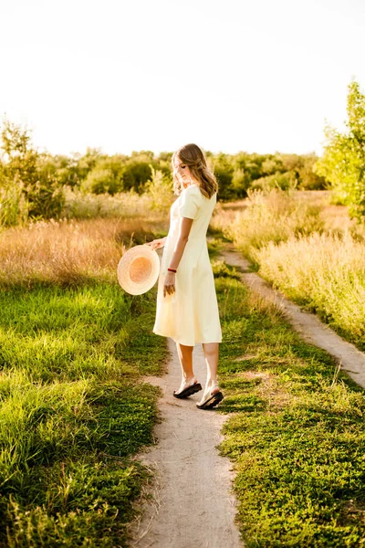 明るい夏の黄色のドレスに金髪の巻き毛を持つ若い美しい女の子と日没時に田舎のフィールドで彼女の手の中にわら帽子 人と自然 — ストック写真