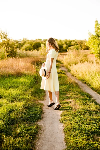 Sarışın, kıvırcık saçlı, açık sarı elbiseli ve elinde hasır bir şapkayla gün batımında kırsaldaki bir tarlada genç bir kız. İnsanlar ve doğa 